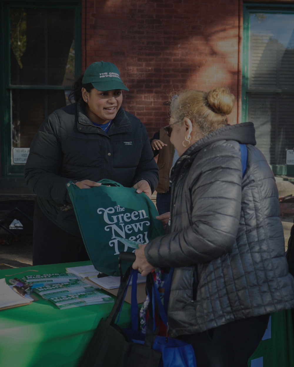 A person in a black puffer jacket and green cap hands a large green tote bag printed with 'to the Green New Deal' to another person standing at an outdoor booth in front of a brick building.